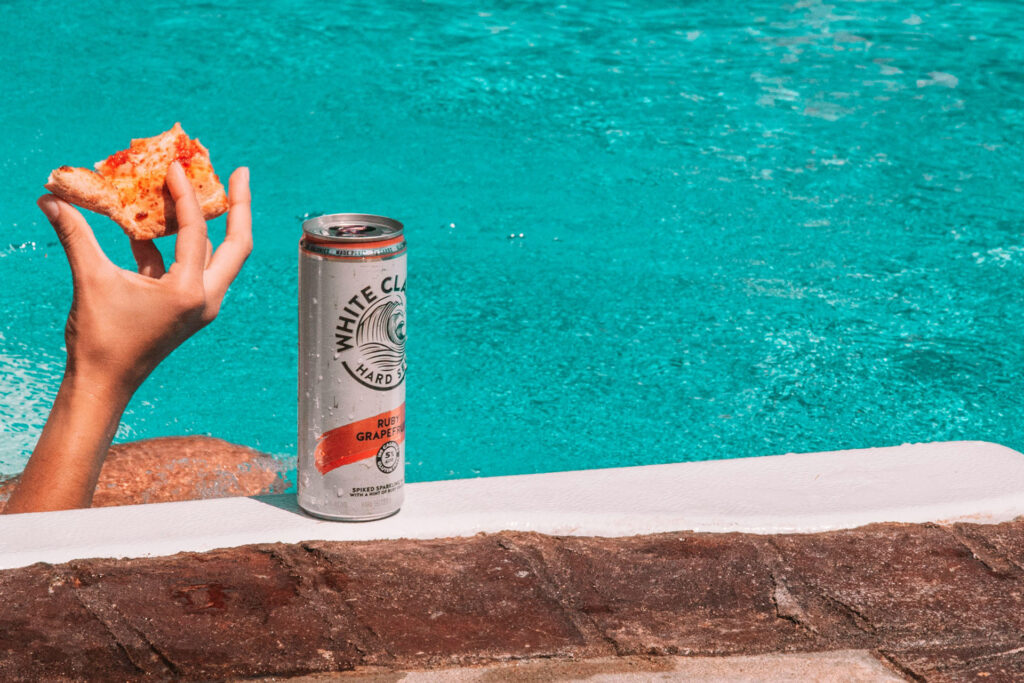 A can of White Claw Ruby Grapefruit hard seltzer RTD sits poolside next to a person holding a slice of pizza, with turquoise water in the background.