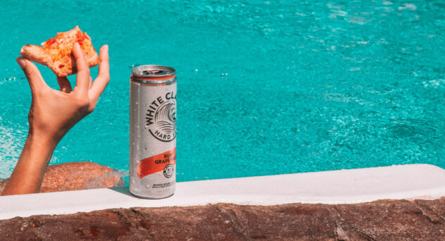 A can of White Claw Ruby Grapefruit hard seltzer RTD sits poolside next to a person holding a slice of pizza, with turquoise water in the background.
